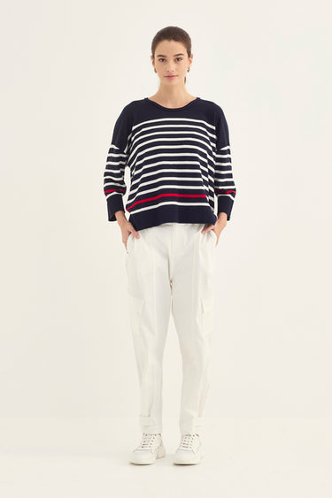 A woman modeling a navy blue knit sweater with horizontal white and red stripes, paired with white trousers by Verge and white sneakers, against a plain off-white studio background.