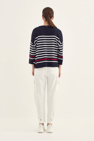 A woman viewed from behind wearing a navy blue sweater with horizontal white and red stripes, paired with white trousers by Verge and white sneakers, against a plain off-white studio background.