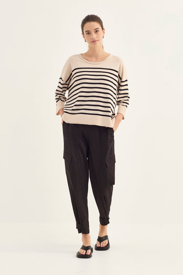 A woman modeling a beige long-sleeve knit top with black horizontal stripes on the sleeves and body, paired with black cargo pants by Verge and black sandals, against a plain white studio background.