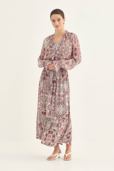 A woman modeling a long-sleeved maxi dress with a dusty rose and beige paisley pattern by Verge, standing against a plain off-white studio background.
