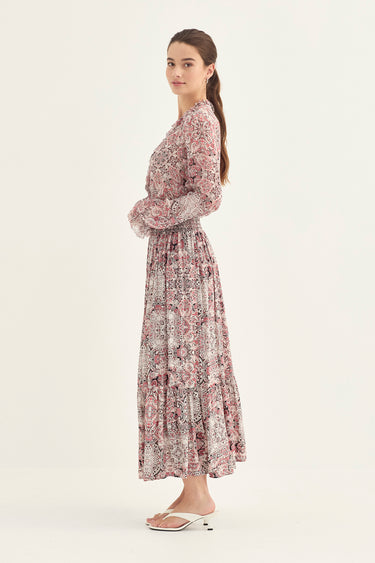 A woman modeling a long-sleeved maxi dress with a dusty rose and beige floral pattern by Verge, standing against a plain off-white studio background.