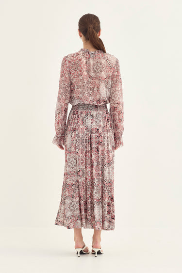 A woman viewed from the back wearing a long-sleeve midi dress with a dusty rose and beige paisley pattern and a matching waist tie by Verge, paired with white heeled sandals against a plain off-white background.