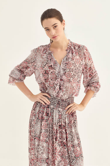 A woman modeling a long-sleeve midi dress with a dusty rose and beige paisley pattern, featuring a ruffled V-neckline and a matching fabric tie belt at the waist by Verge, set against a plain light background.