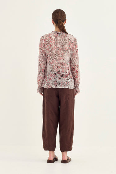 A woman viewed from the back wearing a long-sleeve patterned blouse with a high neck and dark brown cropped pants by Verge, standing against a plain light background.