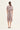 A woman viewed from the back wearing a short-sleeve knee-length dress with an intricate floral and paisley-style print in dusty pink, mauve, and beige tones, paired with white heeled sandals by Verge, against a plain off-white studio background.