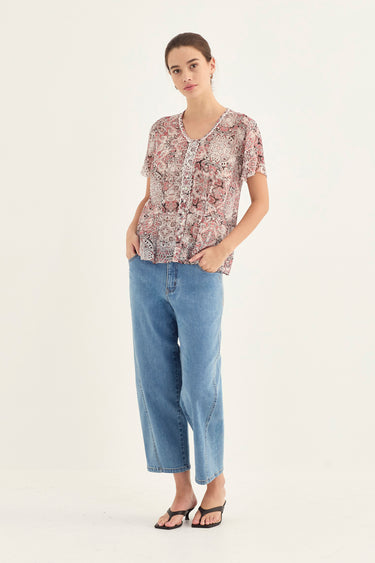 A woman modeling a short-sleeve button-up blouse with a pink, beige, and brown floral pattern, paired with light-wash blue cropped jeans by Verge, and black sandals against a plain white studio background.