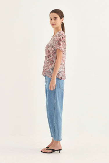 A woman modeling a short-sleeve V-neck top with a pink and beige floral pattern, paired with light blue jeans by Verge and black sandals, shown in profile against a plain white studio background.