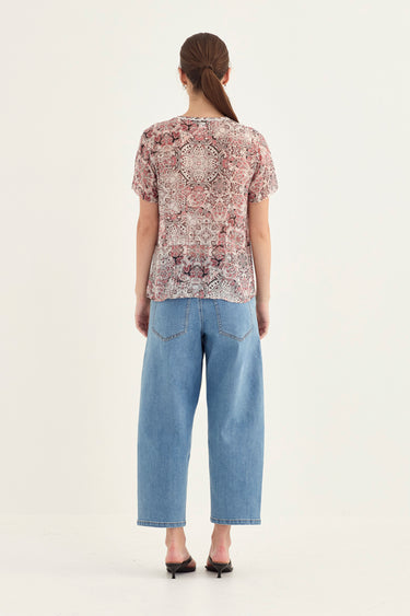 A woman viewed from the back wearing a short-sleeve patterned top with a beige, pink, and brown paisley print, paired with light blue cropped jeans by Verge and black heels, against a plain white studio background.