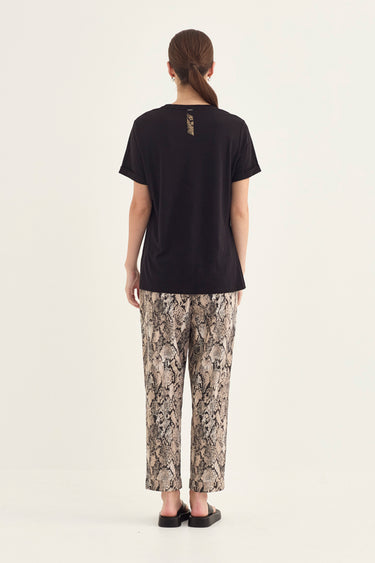 A woman viewed from the back wearing a black short-sleeve t-shirt, paired with beige and brown snakeskin print pants by Verge, against a plain off-white studio background.