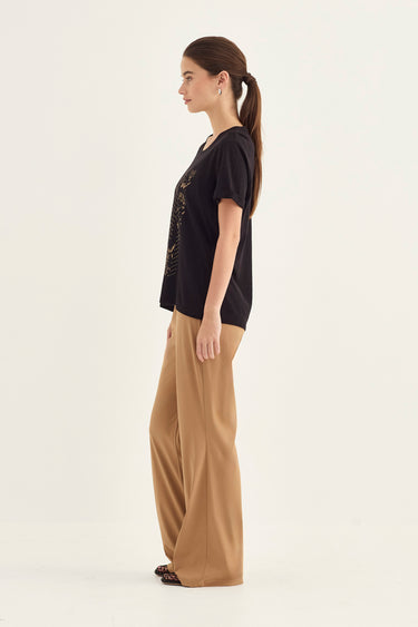 A woman in side profile wearing a black short-sleeve t-shirt with a subtle graphic print and beige wide-leg trousers by Verge, standing against a plain off-white studio background.