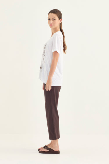 A woman stands in profile wearing a loose white graphic T-shirt, dark brown cropped pants by Verge, and black flip-flop sandals against a plain off-white studio background.
