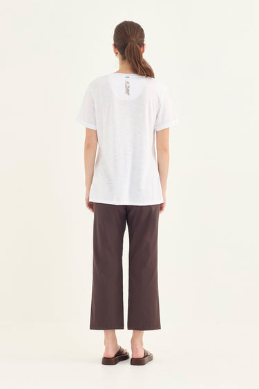 A woman viewed from the back wearing a white short-sleeve top and dark brown cropped pants by Verge, standing against a plain light background.