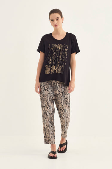 A woman modeling a black short-sleeve t-shirt with a gold-toned abstract graphic print on the front, paired with beige and black snakeskin print pants by Verge and black sandals, against a plain off-white studio background.