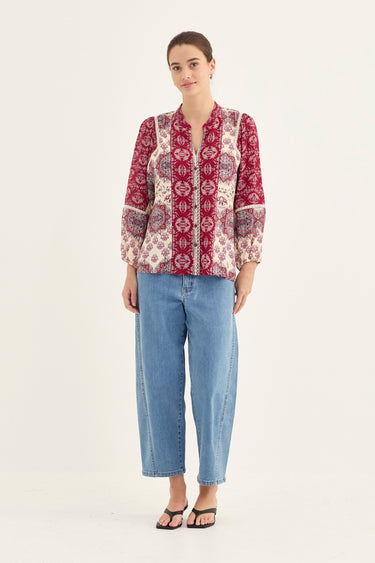 A woman modeling a long-sleeve button-up blouse with a red and beige paisley pattern, paired with light blue cropped jeans by Verge and black sandals, against a plain white studio background.