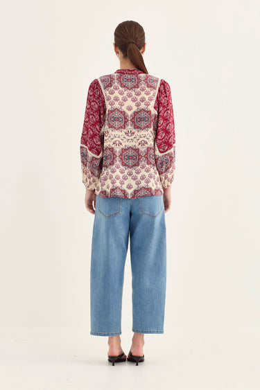 A woman viewed from the back wearing a long-sleeve patterned blouse with a cream base and red paisley sleeves, paired with light blue cropped jeans by Verge and black heels, against a plain light background.