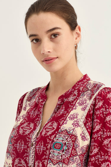 A close-up of a woman wearing a deep red and cream patterned tunic top with intricate embroidery and a V-neckline by Verge, set against a plain light background.