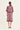 A woman viewed from the back wearing a short-sleeved printed dress by Verge with a cream base and deep red/pink circular floral motifs and border details, set against a plain off-white studio background.
