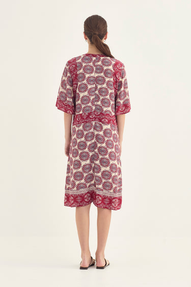 A woman viewed from the back wearing a short-sleeved printed dress by Verge with a cream base and deep red/pink circular floral motifs and border details, set against a plain off-white studio background.