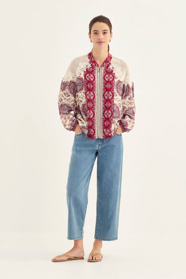 A woman modeling a bohemian-style embroidered bomber jacket with blue jeans by Verge and flat sandals, standing against a plain off-white studio background.