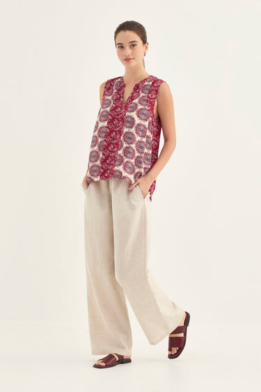 A woman modeling a sleeveless V-neck top with a red and cream paisley pattern, paired with wide-leg beige trousers by Verge and dark brown sandals, against a plain white studio background.