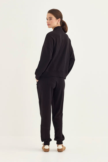 A woman wearing a matching black tracksuit, consisting of a long-sleeve zip-up jacket and jogger pants by Verge, shown from a rear three-quarter view against a plain white studio background.