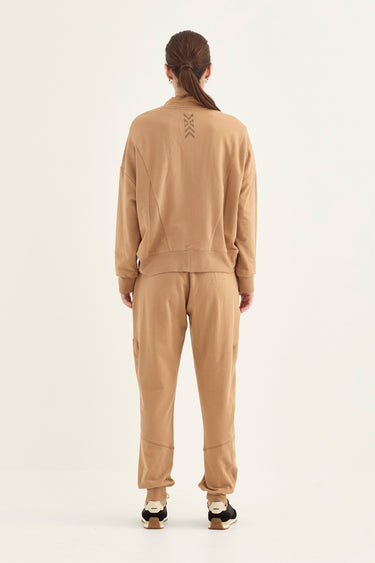 A model viewed from the back wearing a matching tan sweatshirt and jogger pants set by Verge, featuring a small geometric logo on the upper back of the sweatshirt, set against a plain off-white studio background.