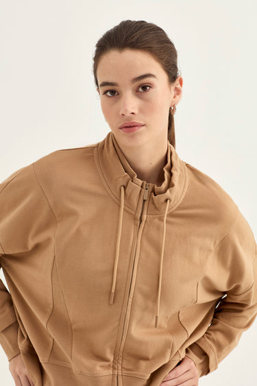 A woman wearing a camel brown zip-up hoodie by Verge with a high mock neck and drawstrings, photographed against a clean off-white studio background.