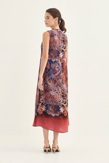 A woman modeling a sleeveless dress by Verge with a complex paisley and floral print in blue, red, and gold, shown from a rear three-quarter view against a plain light background.