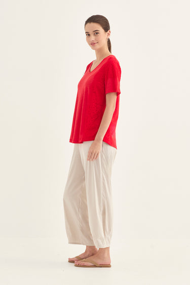 A woman modeling a red short-sleeve V-neck top and cream wide-leg pants by Verge against a plain off-white studio background.