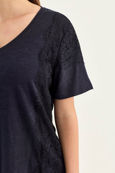 Close-up of a person wearing a dark navy short-sleeve top by Verge with black lace applique panels along the shoulder and side, against a plain light background.