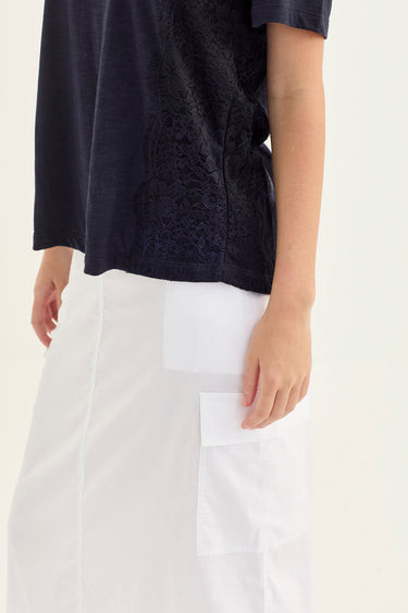 A close-up of a person wearing a dark navy short-sleeve top and a white skirt by Verge with a large front patch pocket, against a plain light background.