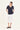 A woman modeling a short-sleeve navy blue V-neck top and a white midi skirt by Verge, paired with black slide sandals, against a plain off-white studio background.
