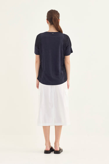 A woman viewed from behind wearing a dark navy short-sleeve t-shirt and a white midi skirt by Verge, standing against a plain off-white studio background.