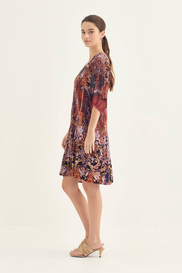 A woman modeling a short-sleeved, knee-length shift dress by Verge with an intricate multicolor paisley and floral print in shades of purple, red, orange, and blue, paired with beige heeled sandals against a plain off-white studio background.