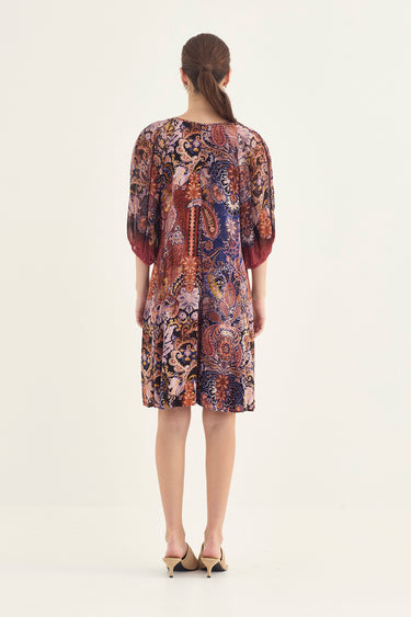 A woman viewed from the back wearing a knee-length patterned dress by Verge with three-quarter sleeves and beige heels, against a plain light background.