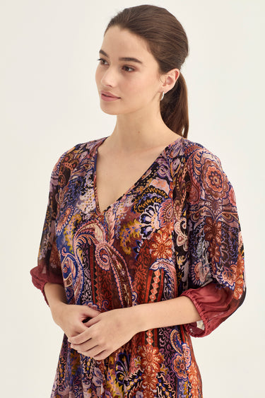 A woman wearing a loose, V-neck dress by Verge with a dense paisley and floral pattern in rust red, blue, purple, and gold tones, photographed against a plain light background.