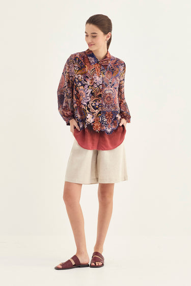 A woman modeling a long-sleeve button-up shirt with a dense multicolor paisley and floral pattern in purple, orange, and blue, layered over a dusty rose undershirt, paired with beige shorts by Verge and dark brown sandals, against a plain off-white studio background.