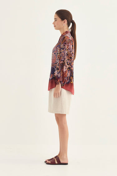 A woman shown in profile wearing a long-sleeve floral print blouse with a deep red hem, paired with an off-white shorts by Verge and dark brown slide sandals, against a plain white studio background.