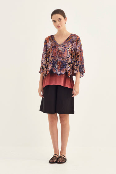 A woman modeling a two-piece outfit consisting of a short-sleeved V-neck top with a multicolor paisley pattern and a dusty rose underlayer by Verge, paired with black shorts and black lace-up sandals, against a plain light background.