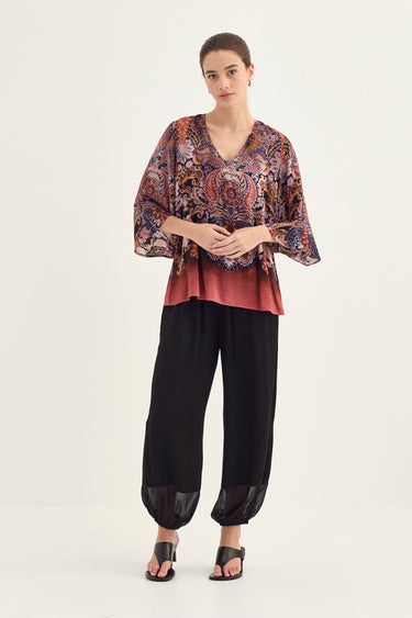 A model stands against a plain light background wearing a loose V-neck top by Verge with a multicolor paisley pattern and a muted red-orange underlayer, paired with black harem-style pants and black sandals.