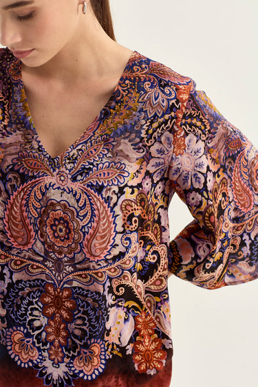 A close-up of a woman wearing a long-sleeve V-neck blouse by Verge  with an intricate paisley and floral pattern in purple, blue, orange, and beige tones.