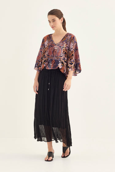 A woman modeling a loose, short-sleeved V-neck top with a colorful paisley and floral pattern in orange, blue, and purple by Verge, paired with a long black pleated skirt and black sandals, against a plain light background.