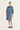 A woman modeling a light blue denim shirt dress by Verge with dark blue embroidered detailing on the chest, styled with black slide sandals against a plain white studio background.