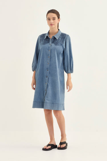A woman modeling a light blue denim shirt dress by Verge with dark blue embroidered detailing on the chest, styled with black slide sandals against a plain white studio background.