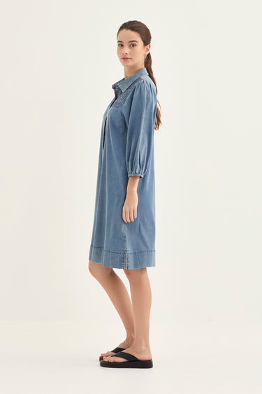A woman modeling a light blue denim shirt dress by Verge with three-quarter puffed sleeves and a relaxed fit, standing in profile against a plain off-white studio background.