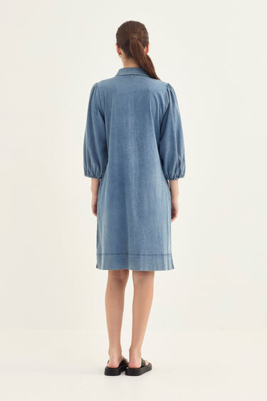 A woman viewed from the back wearing a light blue long-sleeve shirt dress by Verge with gathered sleeves and a straight knee-length hem, standing against a plain off-white studio background.