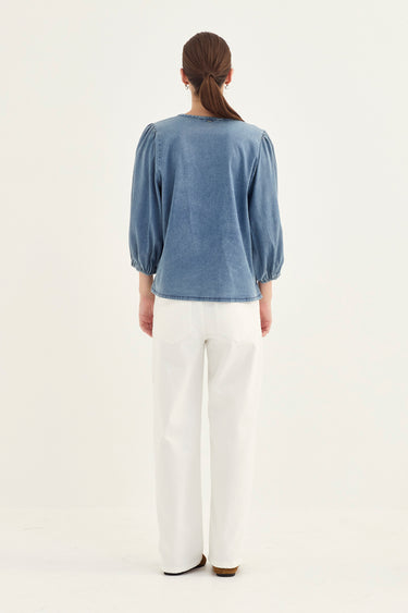 A woman viewed from the back wearing a dusty blue long-sleeve top with puffed shoulders and white wide-leg jeans  by Verge, standing against a plain white studio background.