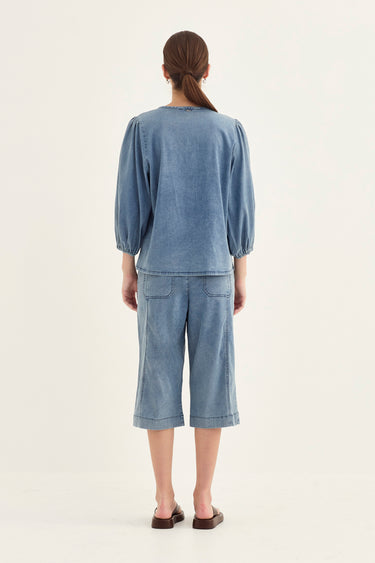A woman viewed from the back wearing a matching dusty blue two-piece set consisting of a long-sleeve top with gathered cuffs and wide-leg cropped pants by Verge, set against a plain off-white studio background.