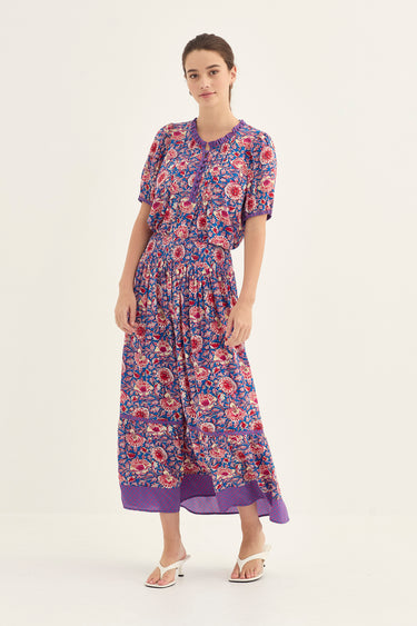 A woman modeling a short-sleeved blouse and a skirt by Verge with a purple, red, and blue floral pattern set against a plain white studio background.