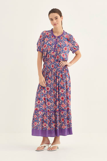 A woman modeling a short-sleeved blouse and a skirt by Verge with a purple, red, and blue floral pattern set against a plain white studio background.